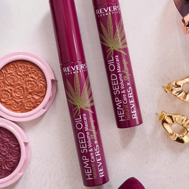 Two Burgundy Hemp Seed Oil mascara tubes on a white background with gold earrings and a pink compact.